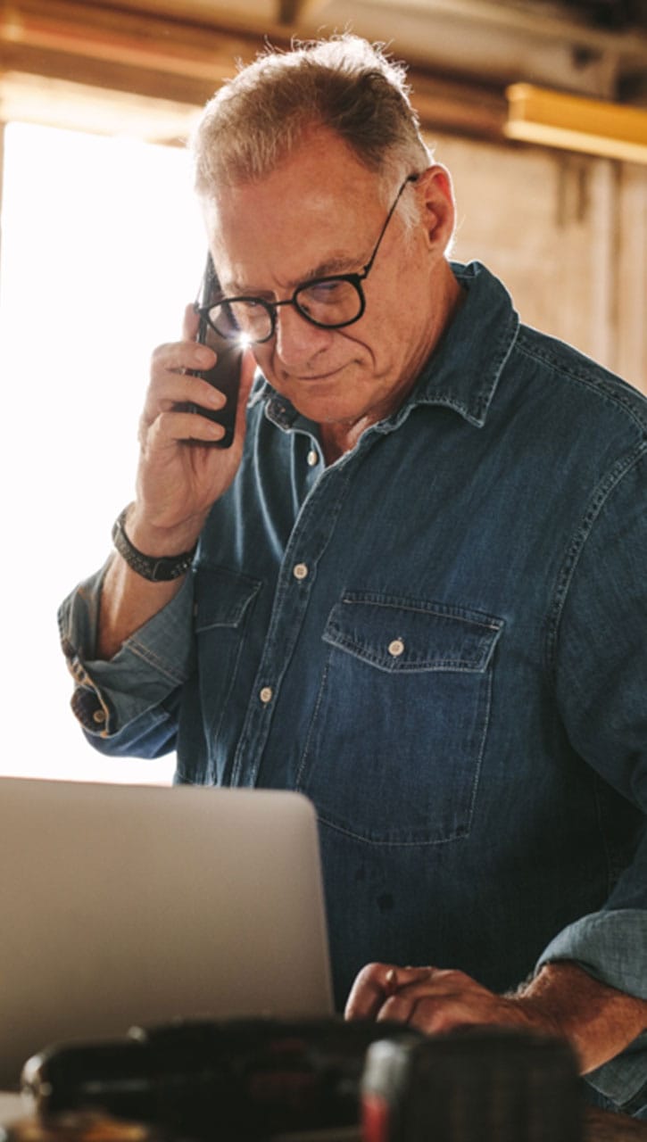 En mann i denimskjorte og briller snakker i telefonen mens han arbeider på sin laptop, med et fokusert uttrykk.